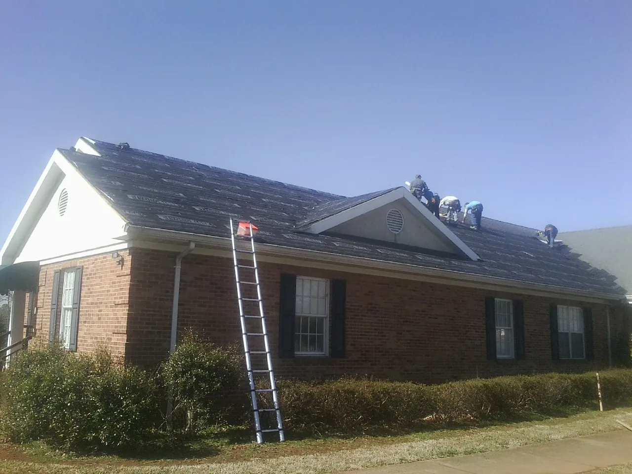 Crew tearing shingles off a brick ranch roof with a ladder set — Emergency Roof Repair work by our crew in Lennox