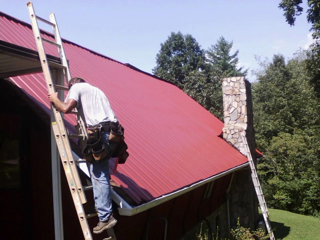Roofer on a ladder beside a red metal roof and stone chimney — Soffit & Fascia Repair by Roofing Brothers in Lennox